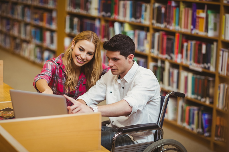 Photo of a young man in a wheelchair sitting at a laptop in a library.  A young woman beside him is pointing to the screen